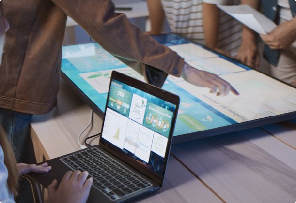 A group of people collaborates around a table, with one person seated at a laptop while others stand around a large interactive screen, manipulating digital charts or maps with their hands.