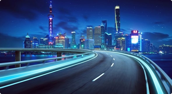 An illuminated city highway at night, winding along a river or bay, with a brightly lit skyline in the background featuring skyscrapers and a prominent tower.