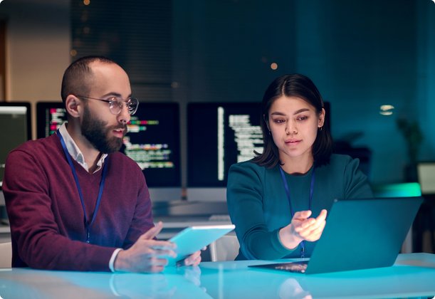 Two developers, a woman and a man, are sitting at a table in front of computer screens displaying code. The woman is pointing at a laptop while the man looks at a tablet.