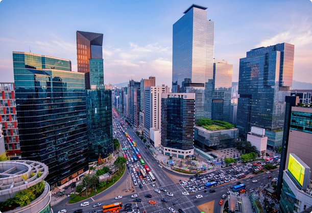An elevated view of a bustling modern city street intersection with numerous cars and pedestrians, surrounded by towering glass skyscrapers and dense urban architecture under a vibrant sky.
