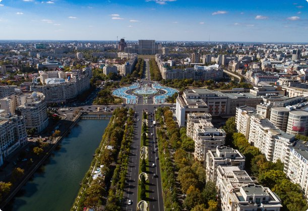 An aerial view of a wide, tree-lined boulevard stretching through a dense city, leading to a large, ornate circular monument or building in the distance, with a river or canal running parallel on the left. The cityscape features numerous residential and commercial buildings under a clear blue sky.