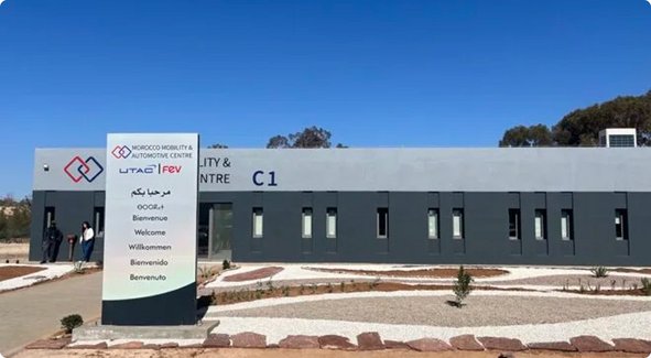 A modern, single-story building with a grey facade and a tall sign displaying multiple company logos and names, under a clear blue sky.