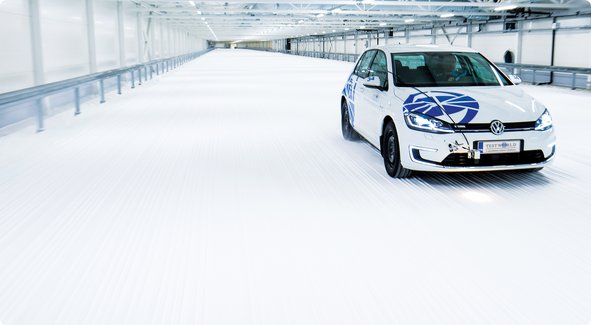 A white car with blue logos is parked in a long, snow-covered testing tunnel or cold-weather facility.