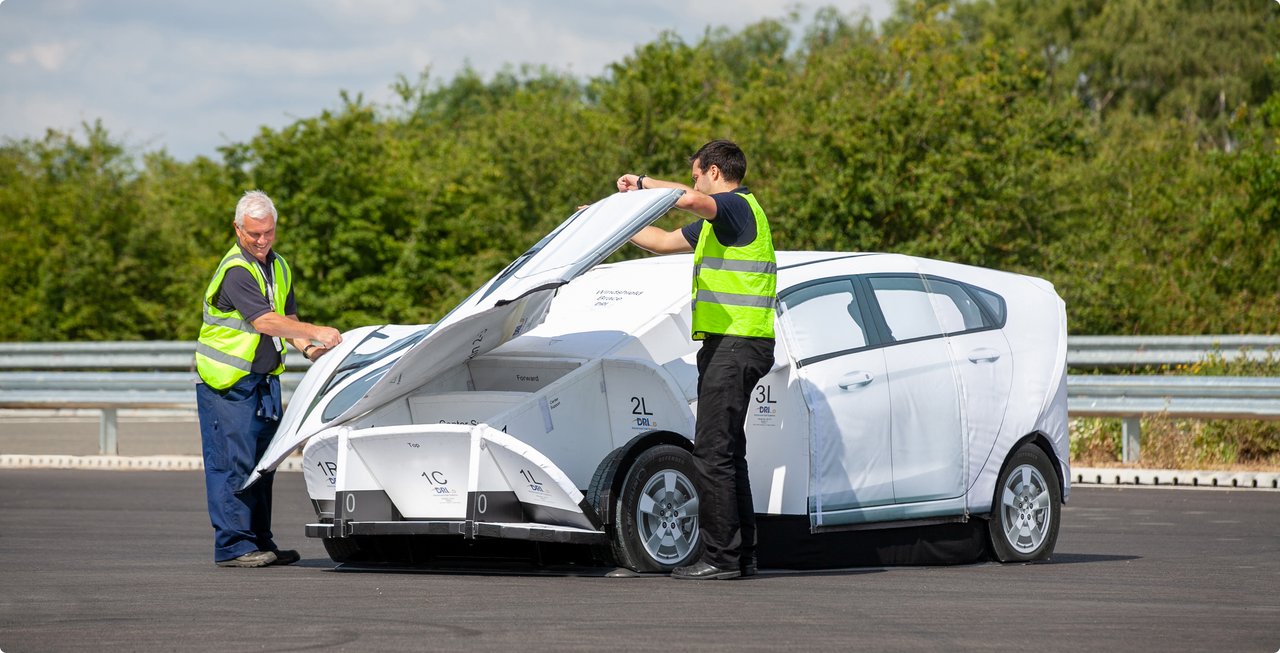 Two employees in high-visibility vests assemble a white, modular covering around a small, white car outdoors, possibly on a test track or road.