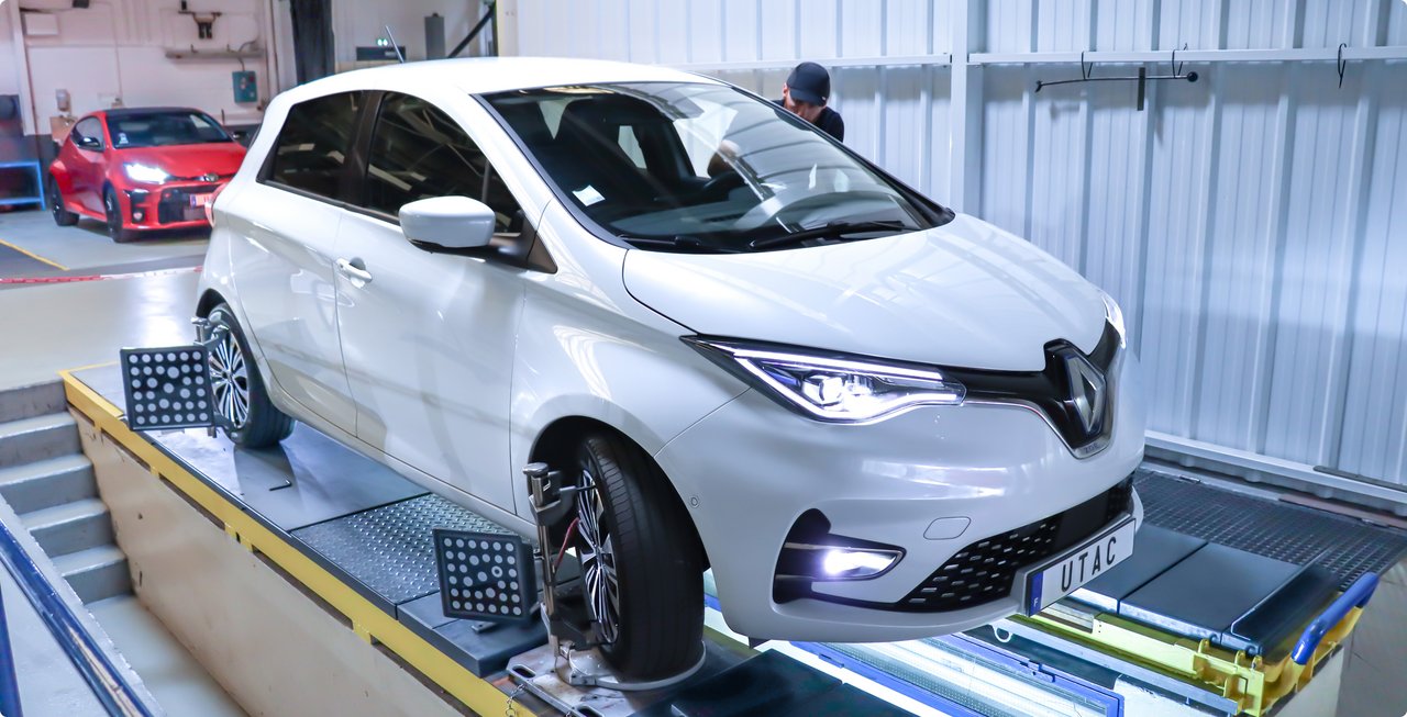 A white Renault Zoe is on a test bench in a workshop or testing center, with a red sports car visible in the background.