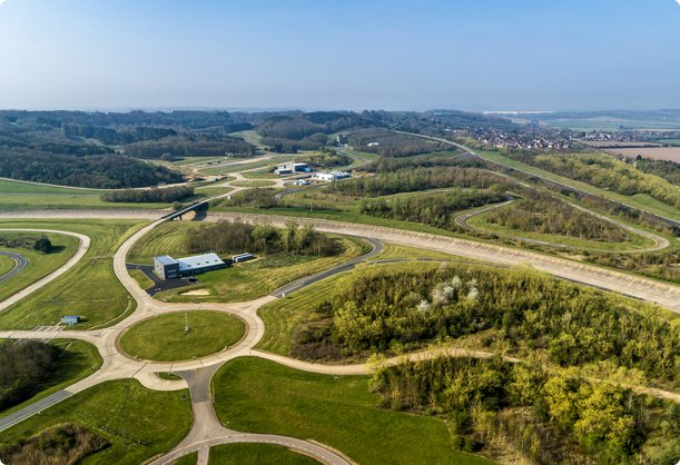 A sprawling aerial view shows a vast, green test track or proving ground, composed of multiple interconnected roads and oval courses, surrounded by forests and open fields under a wide sky.