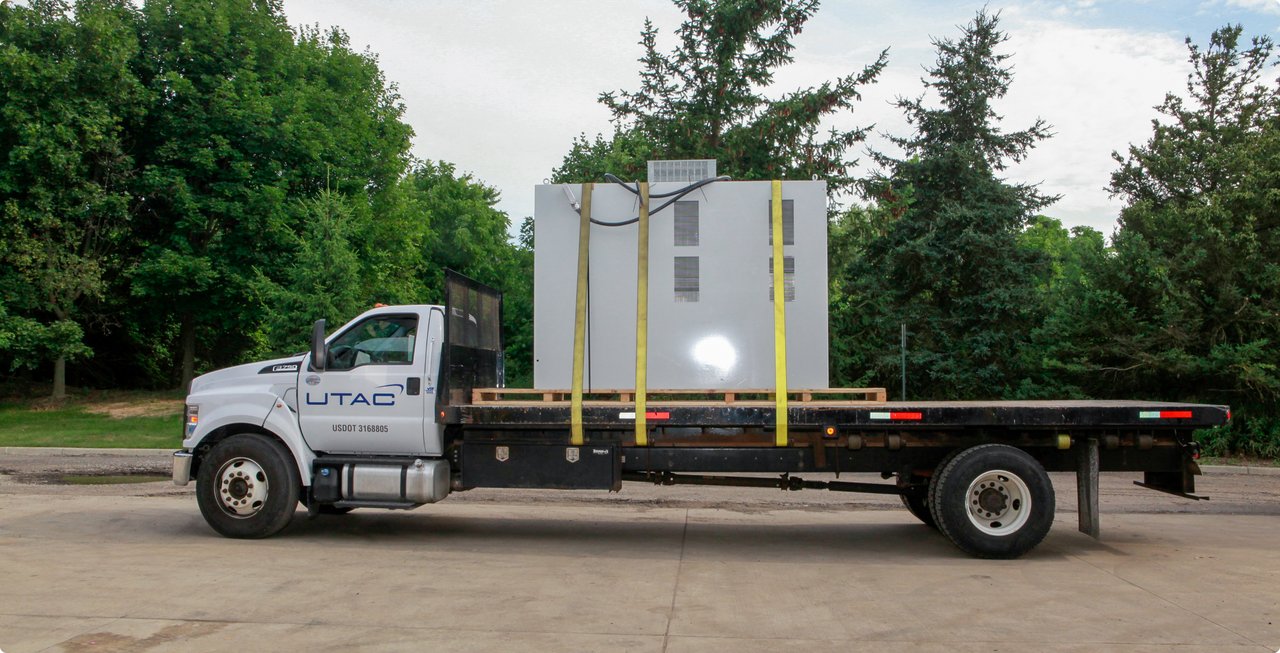 A flatbed truck is parked outdoors, carrying a large, white rectangular object or structure securely fastened with yellow straps. Green trees are visible in the background under a cloudy sky.