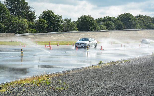 A white car drives on a wet test track marked by cones, with water splashing from its tires.