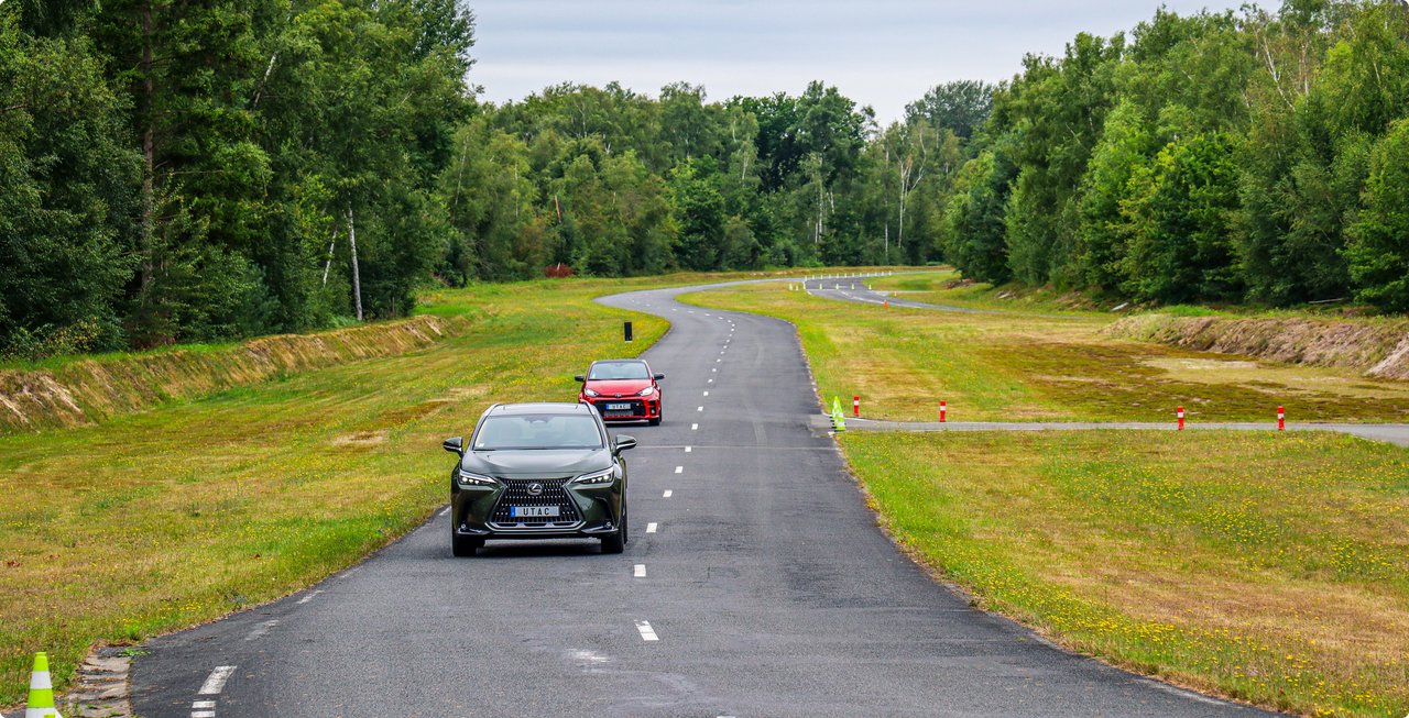 A dark-colored car leads a red car down a winding, narrow paved road surrounded by green trees and grassy fields with wildflowers under a slightly overcast sky.