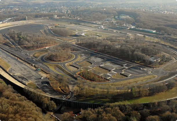An aerial view shows a large test track located in a wooded area with multiple lanes and buildings inside the oval. The trees are bare, suggesting autumn or winter.