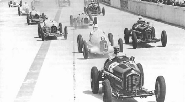A historical black and white image of a line of classic race cars at a starting line, poised for a race.