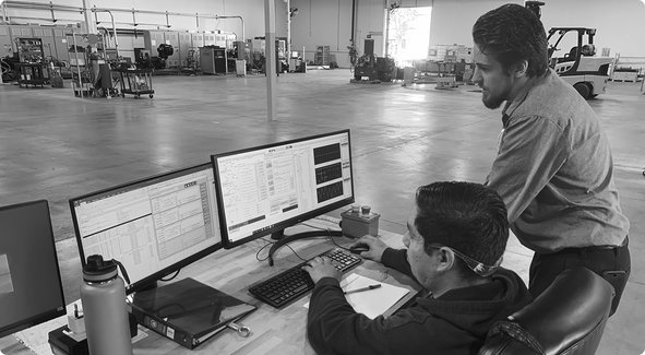 Two individuals observe a computer workstation in a large industrial or warehouse facility with machinery in the background.
