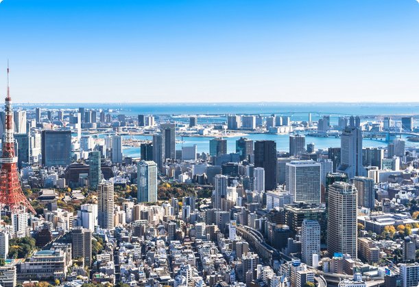 An expansive aerial view of a densely packed city skyline, featuring a prominent red and white transmission tower in the foreground on the left, numerous high-rise buildings, and a large body of water with a distant shoreline under a clear blue sky.