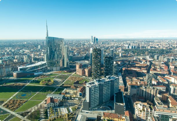 An aerial view of a modern cityscape under a clear blue sky, featuring numerous skyscrapers and high-rise buildings, with a large green park area in the foreground. The city extends towards the horizon with a mix of contemporary and traditional architecture.