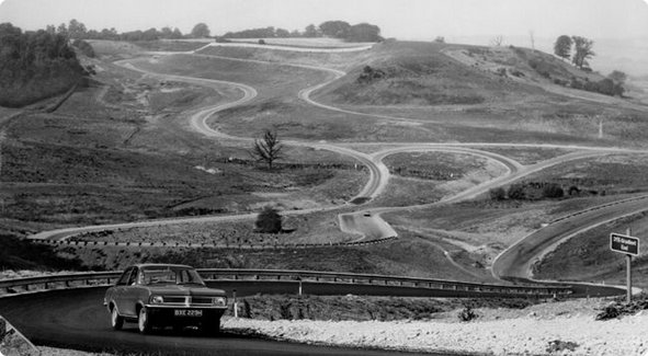 A historical black and white image of a hilly, winding race track or test track with a car driving in the foreground and other tracks extending across the landscape.