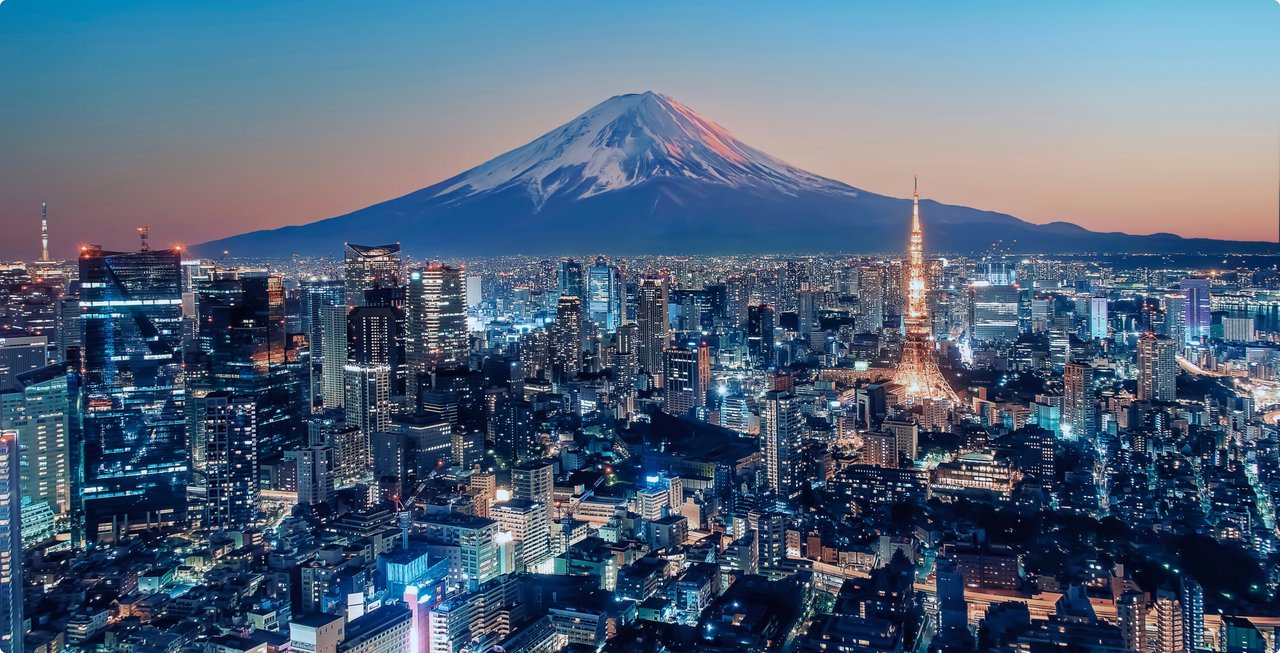 An impressive night view of a densely populated city with countless bright lights, stretching out beneath a large, snow-capped mountain. The sky in the background shows the last colors of twilight.