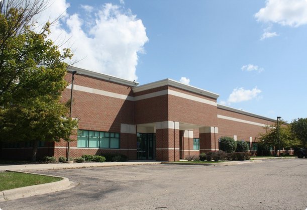 A low-angle exterior view of a modern brick building with large windows and a subtle green tint, surrounded by some trees and a gravel parking area under a partly cloudy sky.