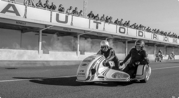 A historical black and white image of a sidecar motorcycle on a race track, with spectators in a grandstand in the background and the word "AUTODROM" visible.