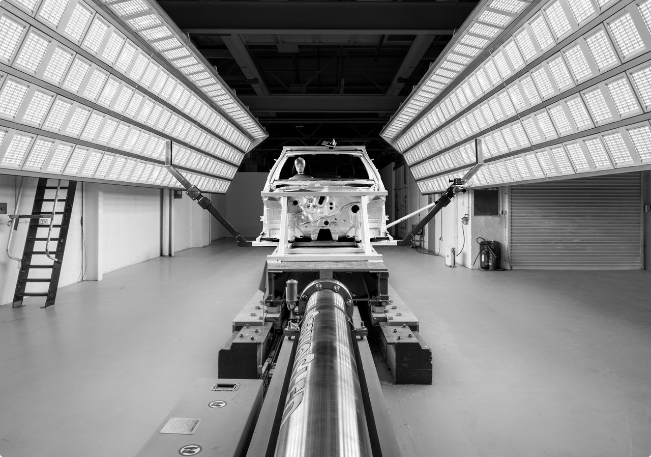 An empty car body on a test stand in a brightly lit testing room with rows of lights on the sides.