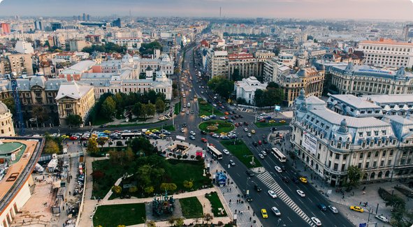 An aerial view of a bustling European city with wide boulevards, historical and modern buildings, heavy traffic, and a large square or park in the center.
